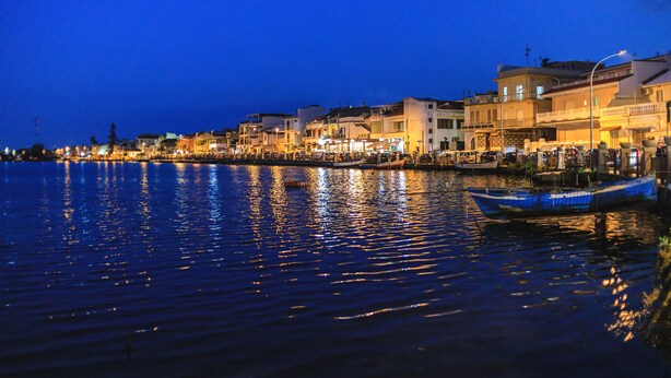 Un paisaje urbano frente al agua de noche, con edificios a lo largo de la costa, un muelle y un bote, además de reflejos en el agua.