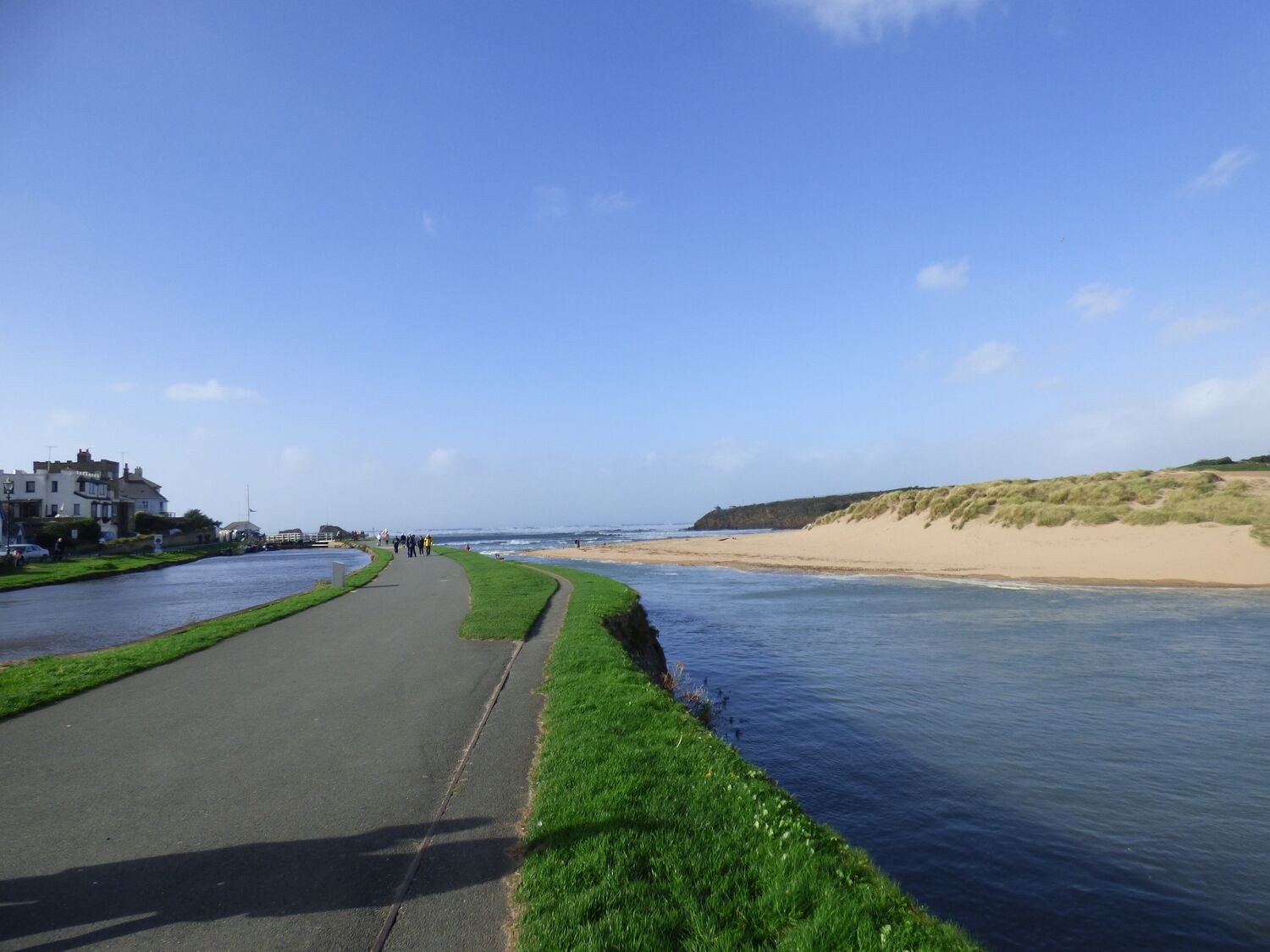 Une route côtière bordée d’un sentier herbeux longeant une rivière, qui mène à une plage de sable.