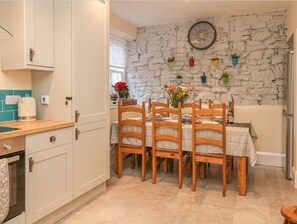Une cuisine avec des meubles blancs, une table à manger en bois avec des chaises, un mur en pierre orné de plantes suspendues et une horloge.