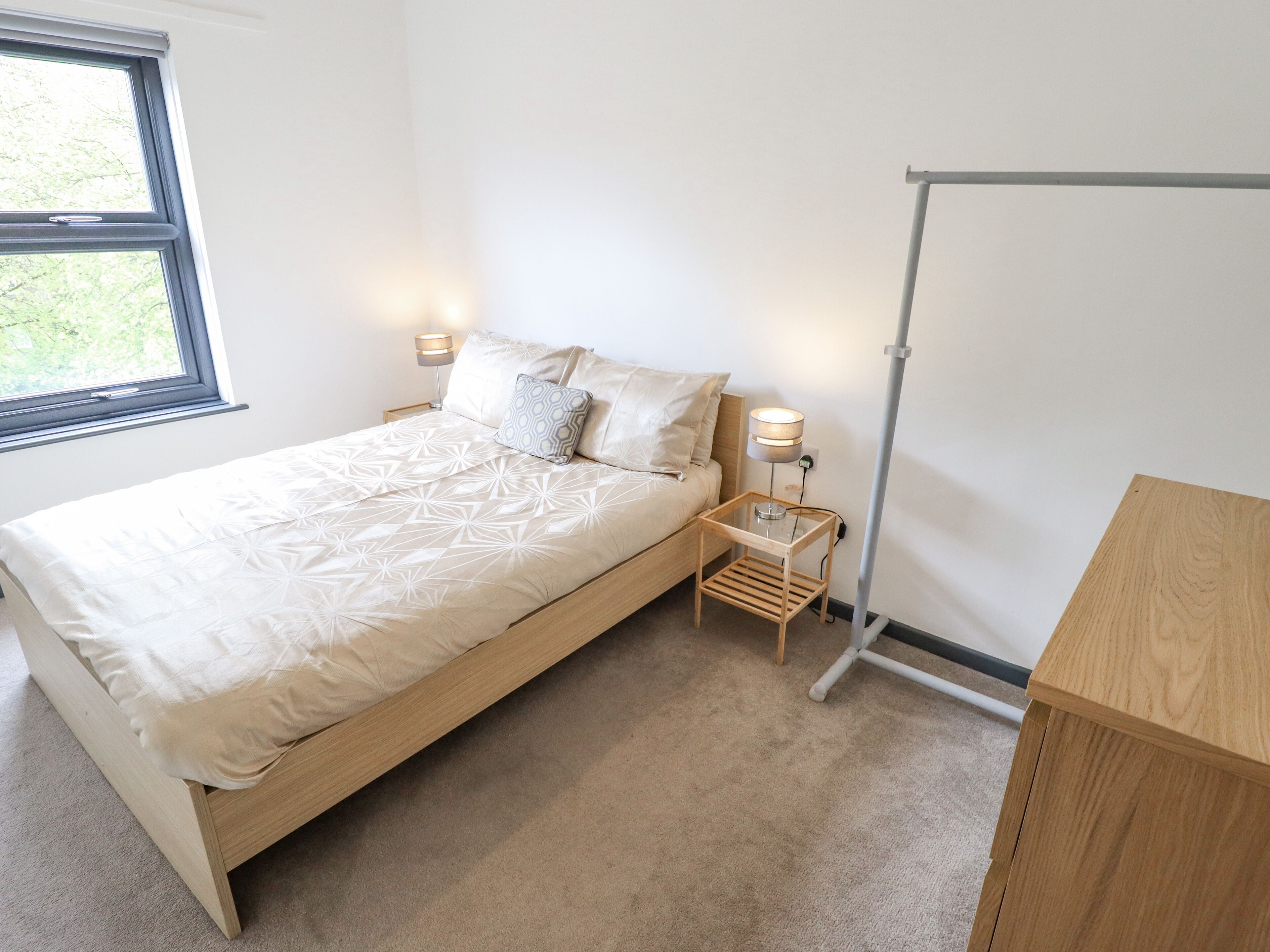 A bedroom with a bed, bedside lamps, a wooden nightstand, a window, and a wooden desk.