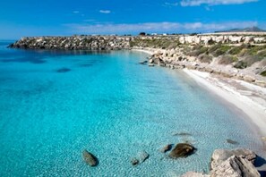 A coastal beach with clear turquoise waters, rocky shoreline, and a distant rocky outcrop.