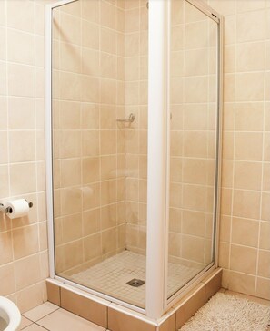 Lasiseinöillä varustettu suihkutila, jossa on beigenväriset laatat, valkoinen lattia ja valkoinen matto alustassa.