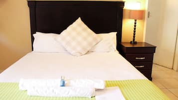 A neatly made bed with white linens and a checkered pillow, a bedside lamp, a small bottle, a folded towel, and a piece of paper on a green-striped bedspread.