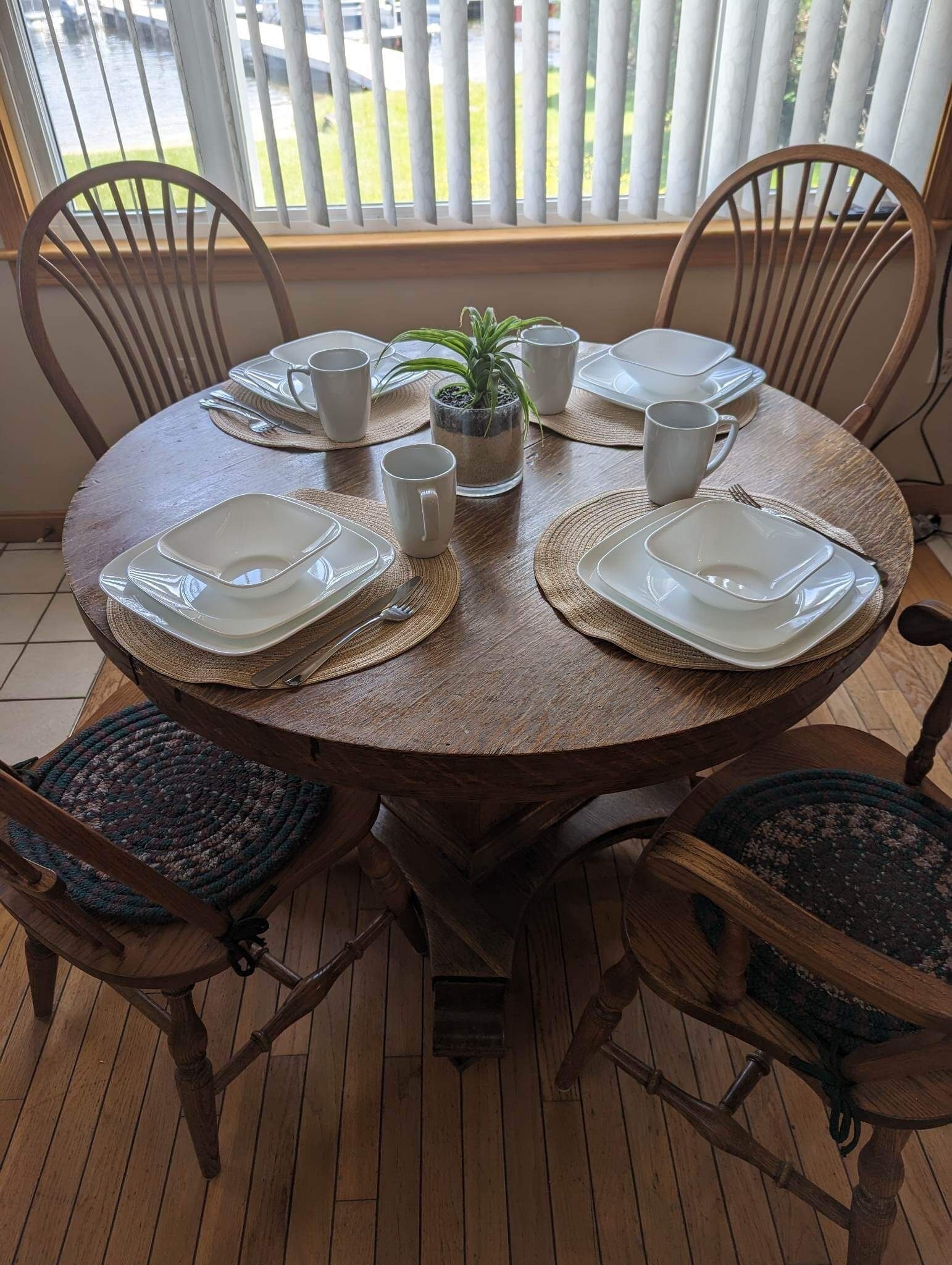 A round wooden dining table with white square plates, clear glass cups, and a small potted plant.