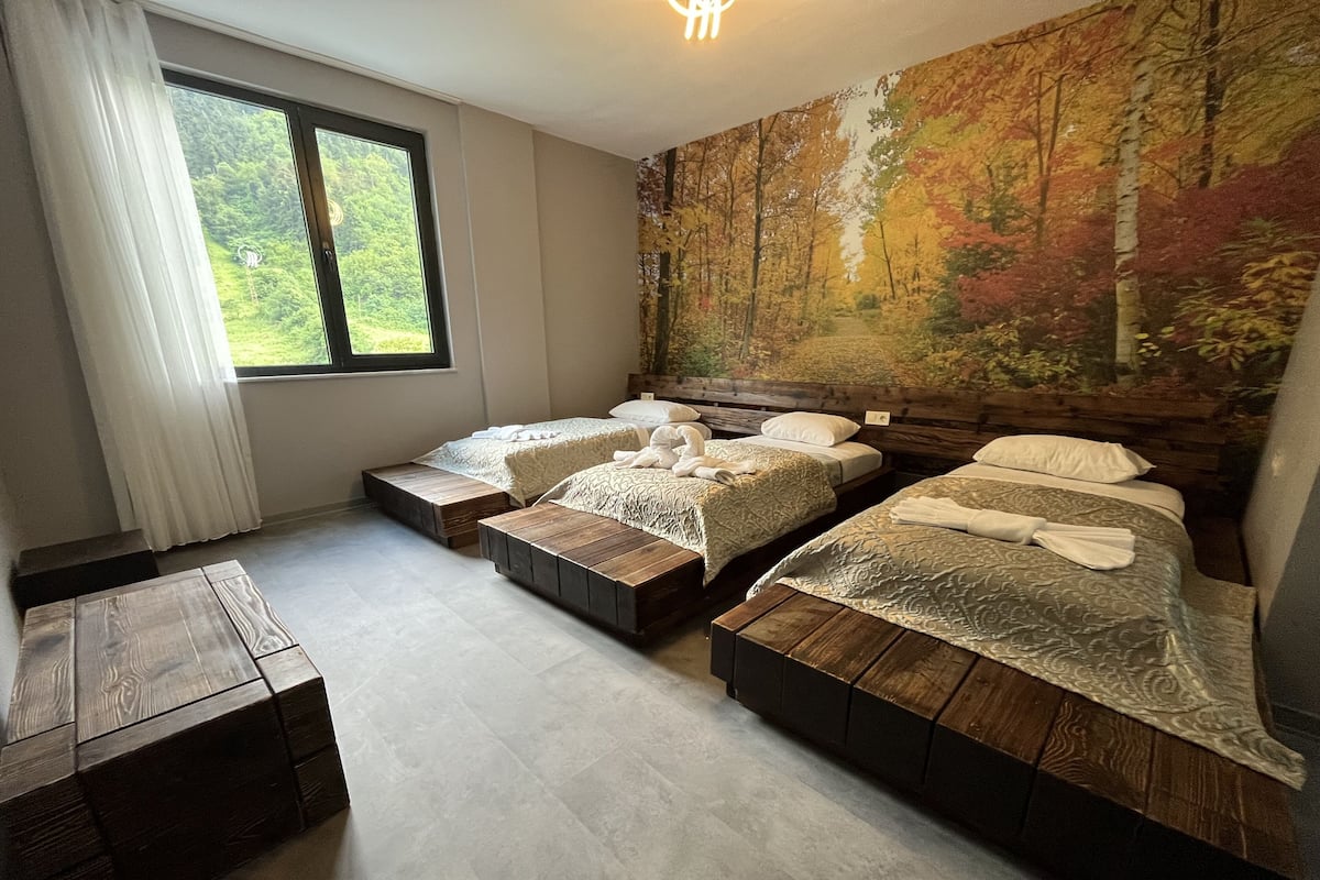 A bedroom with two beds, a wooden bench, and a large window with a view of greenery.