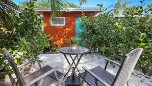 Une terrasse avec deux chaises en métal et une petite table ronde, entourée d’une végétation luxuriante et d’un bâtiment à la façade rouge et turquoise.