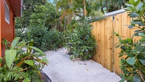 Un étroit sentier en béton, bordé d’une végétation luxuriante et d’une clôture en bois, mène à un bâtiment à la façade rouge.