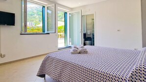 A bedroom with a bed, a TV mounted on the wall, and a bathroom visible through an open door. - Villa in Lemon Grove With Sea Views & Sea Breeze (Minori)