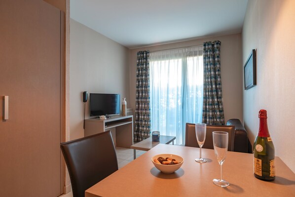 Een hotelkamer met een eettafel, twee stoelen, een schaal met snacks, twee wijnglazen, een fles wijn, een televisie en een raam met gordijnen.