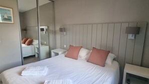 A hotel room with a large bed, a bedside table, a nightstand, a mirror, and a wall-mounted light fixture.