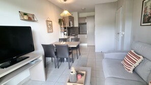 A modern living room with a flat-screen TV, a sofa, a dining area with chairs, and a kitchen in the background.