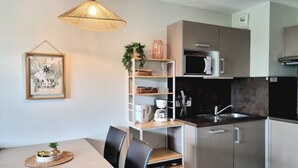A modern kitchen with a microwave, coffee maker, and a sink. There is a dining table with chairs and a hanging light fixture. - Les Sables 504 - Appt avec piscine partagée (Concarneau)