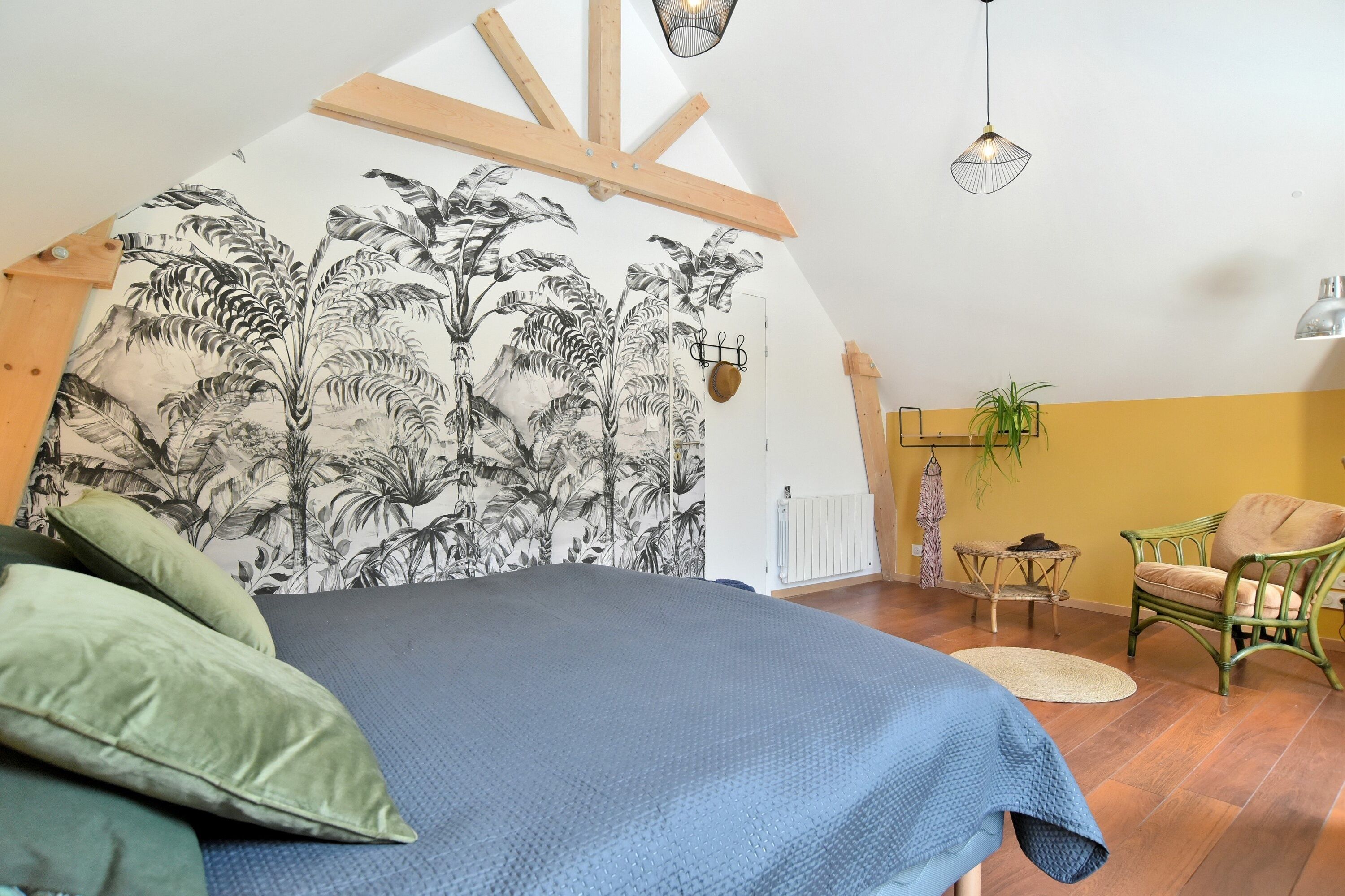 Ein Schlafzimmer mit einem Bett, einer Wand mit tropischem Blattmuster, einem grünen Sessel, einem kleinen Tisch und einer Pendelleuchte.