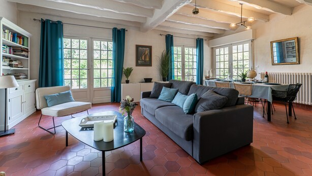 Ein geräumiges Wohnzimmer mit grauem Sofa, einem weißen Sessel und einem Essbereich mit Tisch und Stühlen. Ein Bücherregal voller Bücher, eine Vase mit Blumen und ein Fenster mit blauen Vorhängen sind ebenfalls vorhanden.