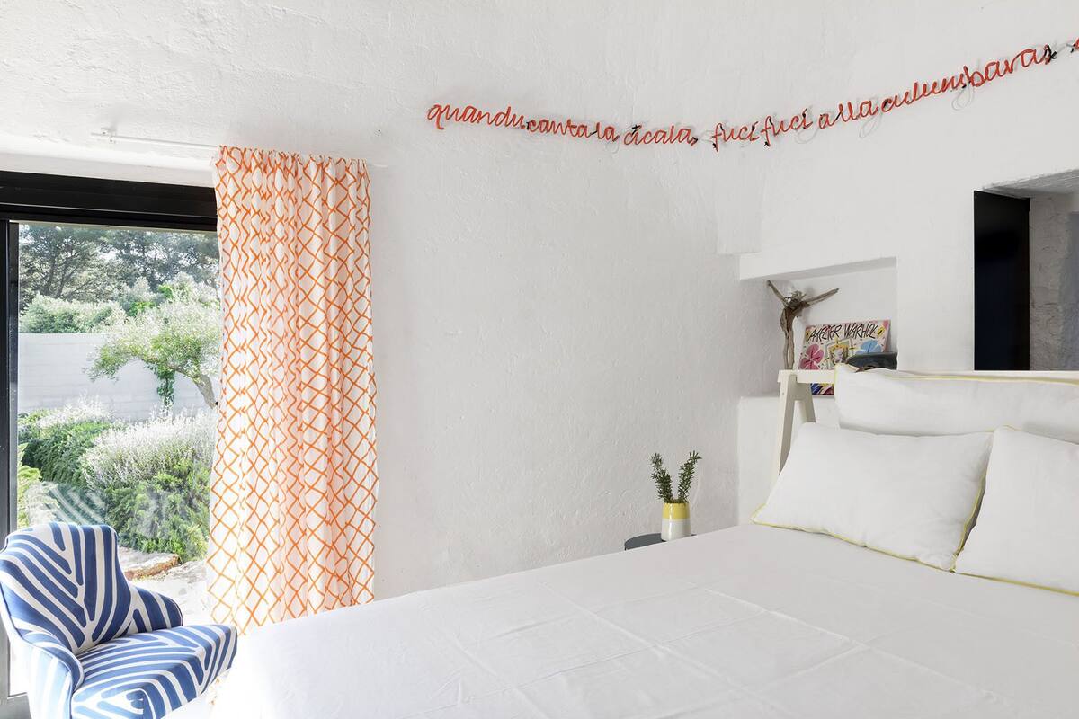A bedroom with a bed, a chair, a small table with a plant, and a wall with a decorative item.
