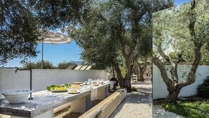 Un espacio de comedor al aire libre con una mesa larga dispuesta para una comida, rodeado de olivos y una pared blanca.