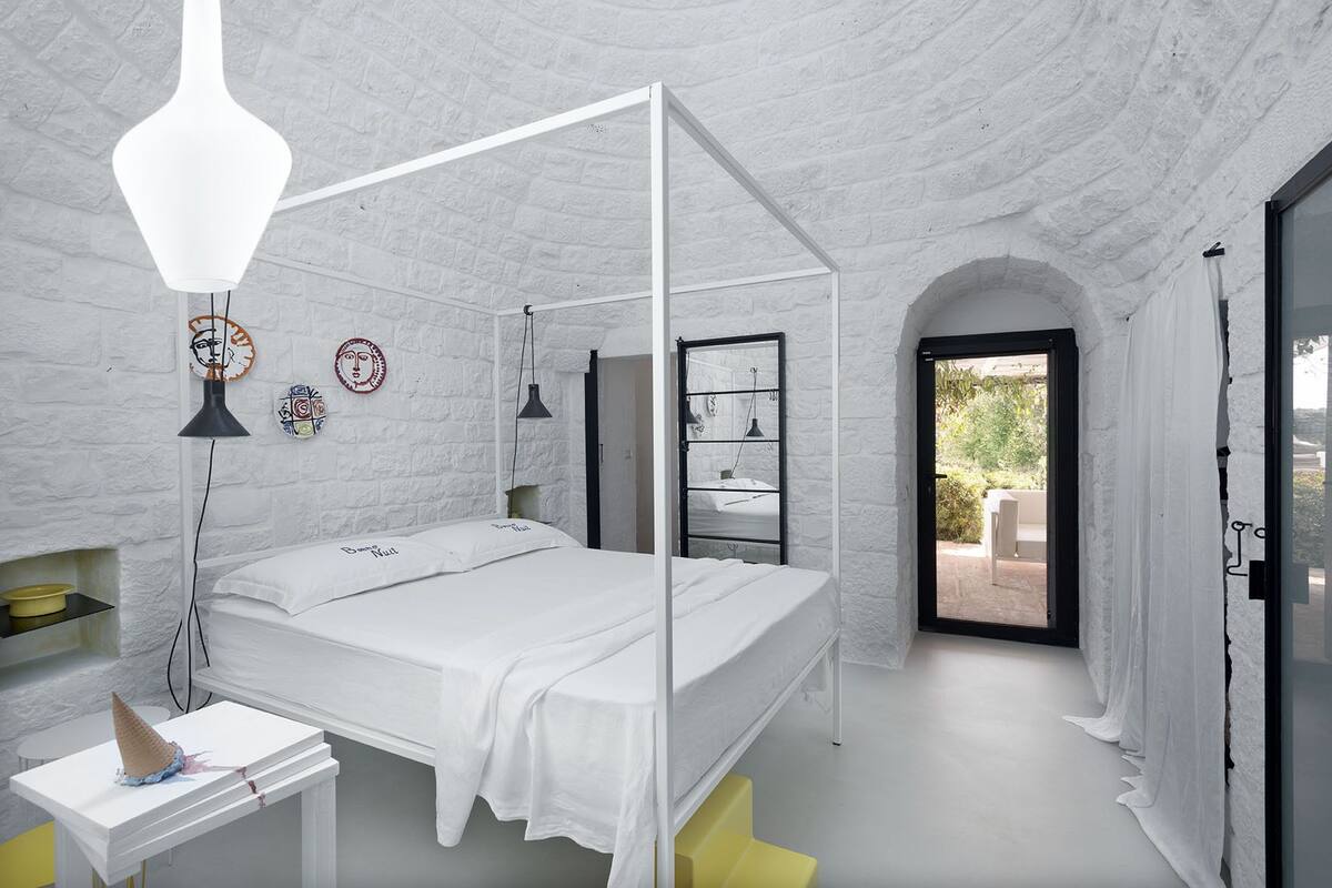 A bedroom with a four-poster bed, a bedside table, a lamp, and a view of the outdoors.