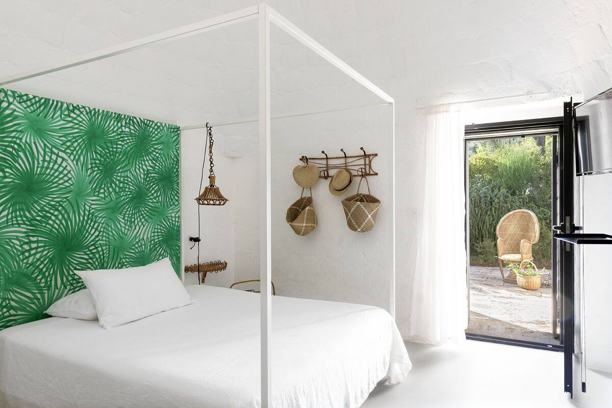 A modern bedroom with a four-poster bed, a green leafy wall, a hanging light fixture, and a wicker basket wall hanging.