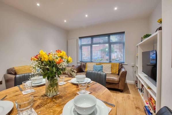 A dining area with a wooden table set for four, a vase of flowers, and a sofa with cushions.