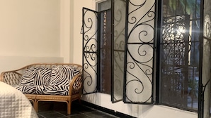 Una habitación con un amplio ventanal con celosías metálicas decorativas, un sofá de mimbre con cojines estampados y suelo de baldosas.