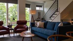A modern living room with a blue sofa, leather chairs, a small table with a book, a floor lamp, and a staircase. - Melrose Townhome Balcony|Walkable to Coffee & Eats (Berry Hill)