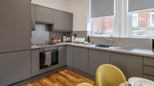 A modern kitchen with grey cabinets, a built-in oven, a sink, and a window with a view of a brick building. - Routh Retreat (Whitby)