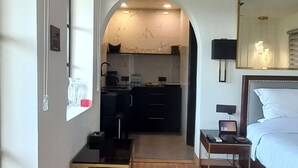 Habitación de hotel con suelo de madera, un escritorio, una cama, una mesita de noche, un espejo y una zona de cocina visible a través de un arco.