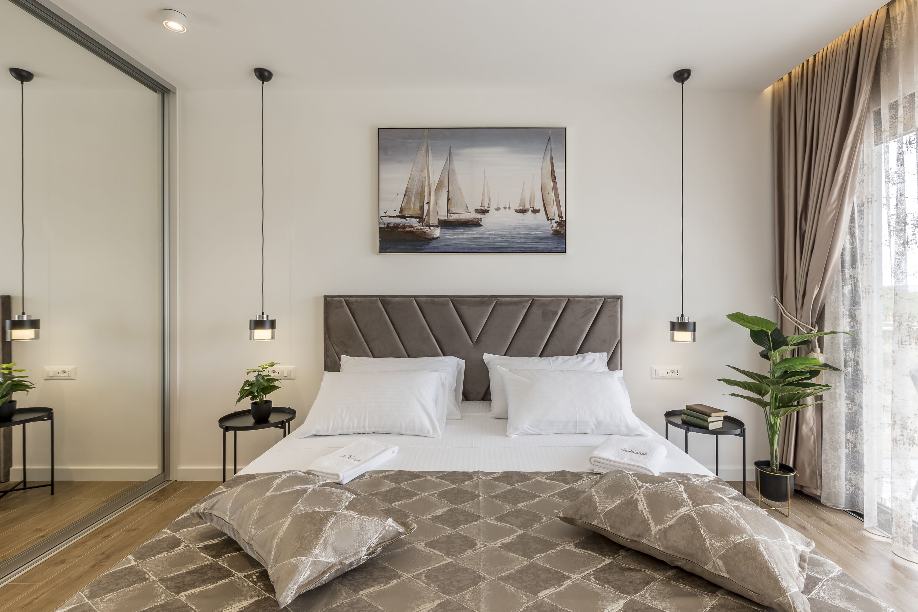 A modern hotel room with a large bed, a headboard, a painting of sailboats, bedside tables with lamps, and a potted plant.
