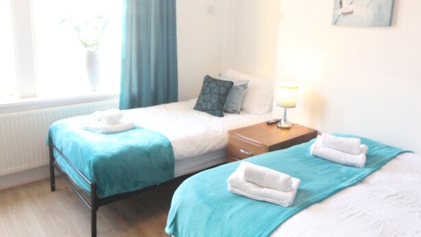 A hotel room with two beds, a nightstand, a lamp, and a window with curtains. - Lundwood House (Barnsley)