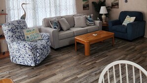 A cozy living room with a ceiling fan, a wooden floor, a sofa, a blue armchair, a coffee table, and a chair with a cushion. - Comfortable cottage in gated community, 5 mins to OC and Assateague (Berlin)