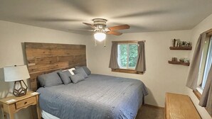 Ein Schlafzimmer mit Bett, Nachttisch, Deckenventilator, Fenster mit Vorhängen und einem Holz-Kopfteil.