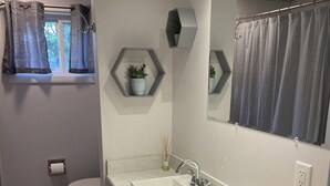 Ein Badezimmer mit einem weißen Waschbecken, einem Spiegel, einer Toilette und einem Fenster mit Vorhängen. Es gibt zwei sechseckige Ablagen mit Pflanzen und ein kleines Fenster mit Vorhang.
