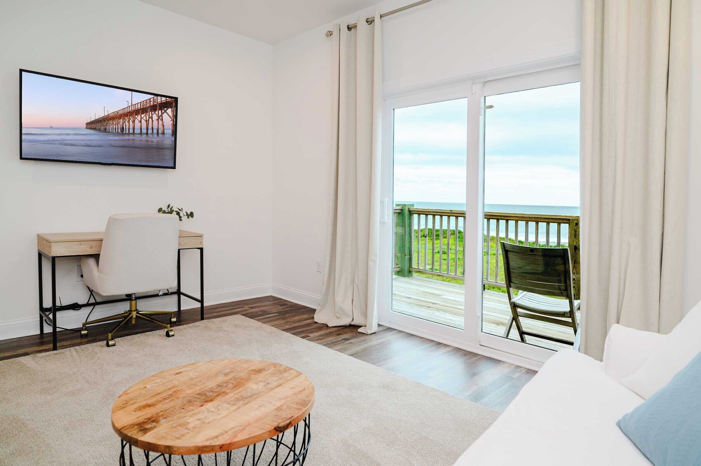 A room with a wooden desk, a white chair, a round wooden table, and a large window with a view of a balcony and a pier.