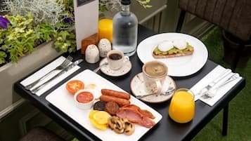 A breakfast table with eggs, sausages, tomatoes, mushrooms, and ham. There are also two glasses of orange juice and a bottle of water.