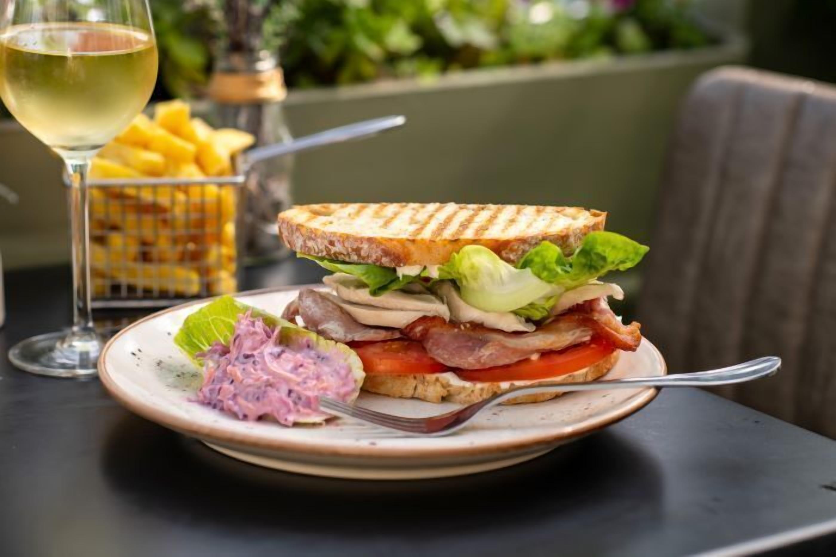 Un sandwich garni de laitue, de tomates et de viande, accompagné d’un verre de vin blanc et d’une portion de frites.