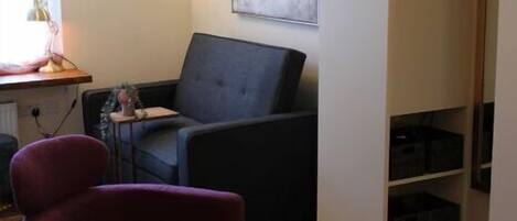 A hotel room with a dark grey sofa, a purple armchair, a small side table with a lamp, a large abstract painting on the wall, and a built-in shelving unit with storage boxes.
