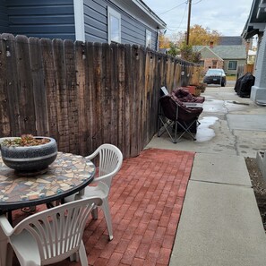 Eine Terrasse mit rundem Tisch, zwei weißen Stühlen, einem Blumentopf und einem Pflasterweg.