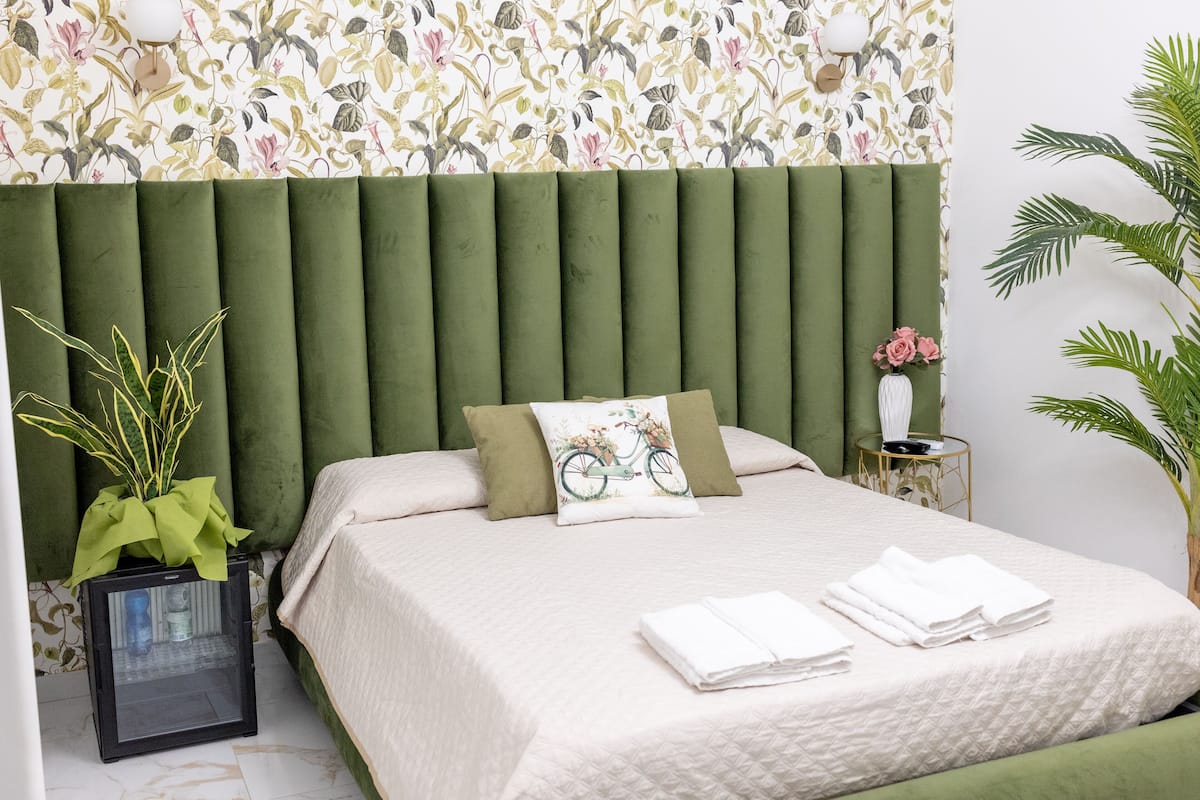 Cama bem arrumada com cabeceira verde, uma pequena planta em vaso e parede com papel de parede floral.