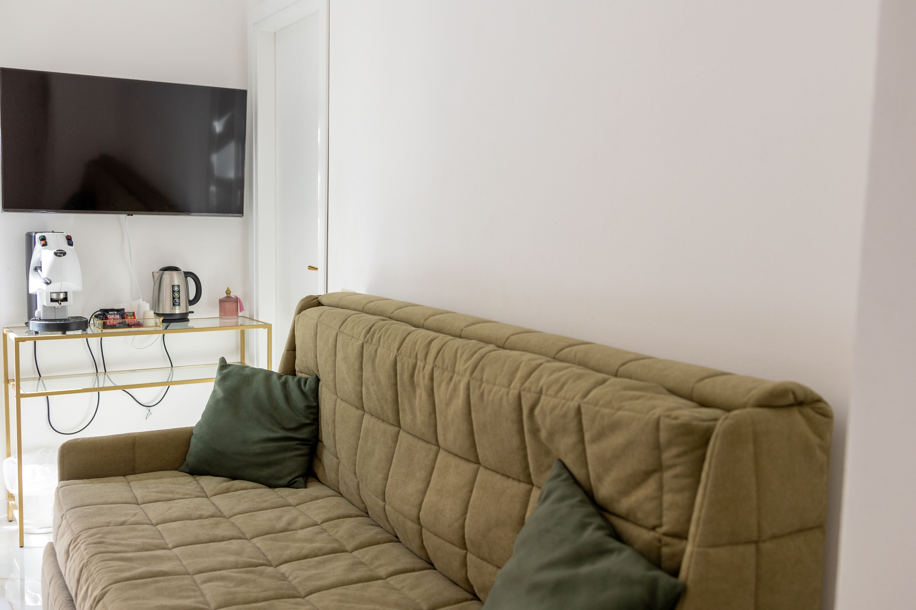 Ein beiges Sofa mit einer gesteppten Rückenlehne, ein Flachbildfernseher an der Wand montiert und eine Kaffeemaschine auf einem Glastisch neben dem Sofa.