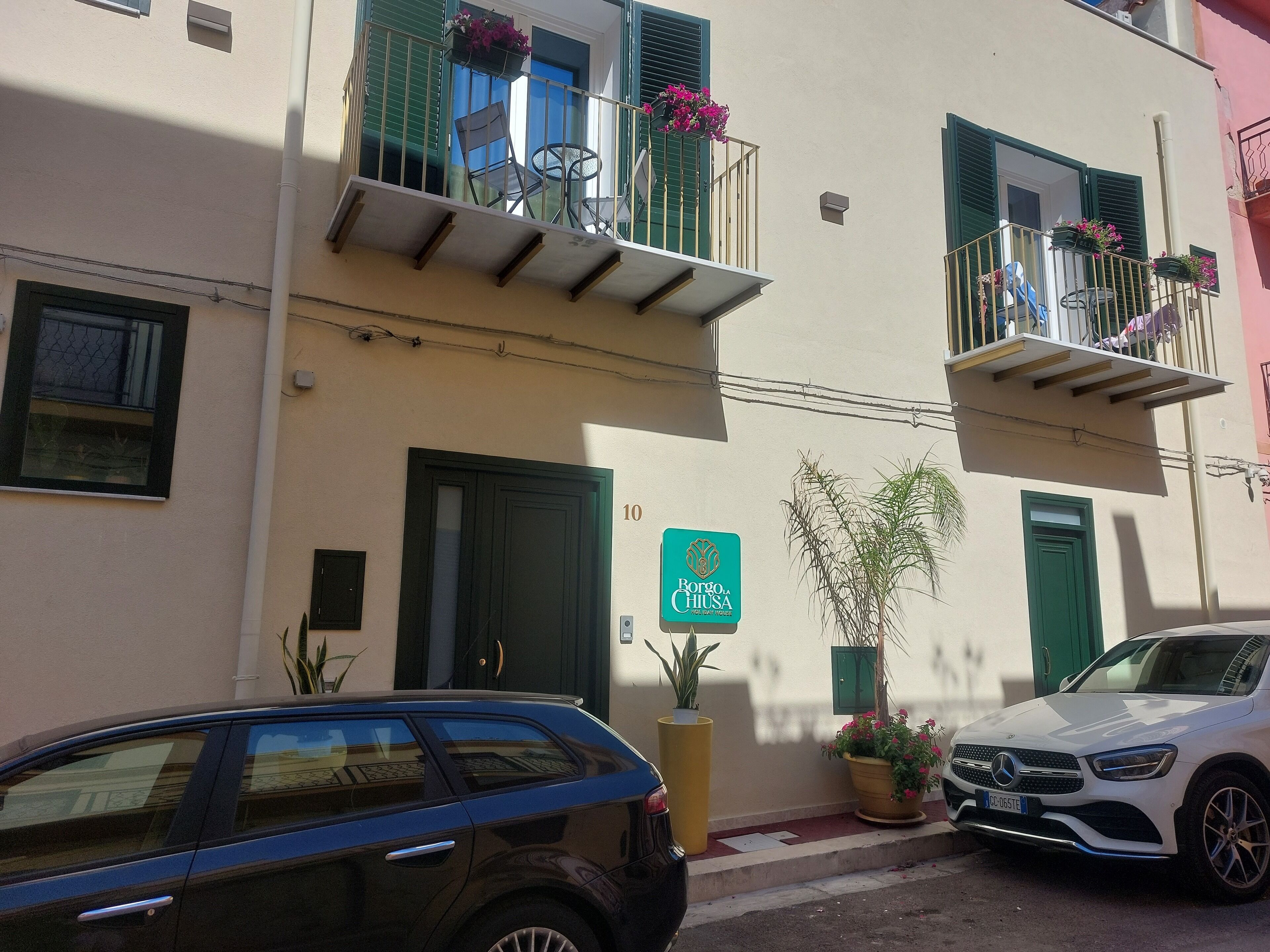 Un edificio con un cartello che indica 'Borgo Chiusa', balconi adornati con piante in vaso e auto parcheggiate.