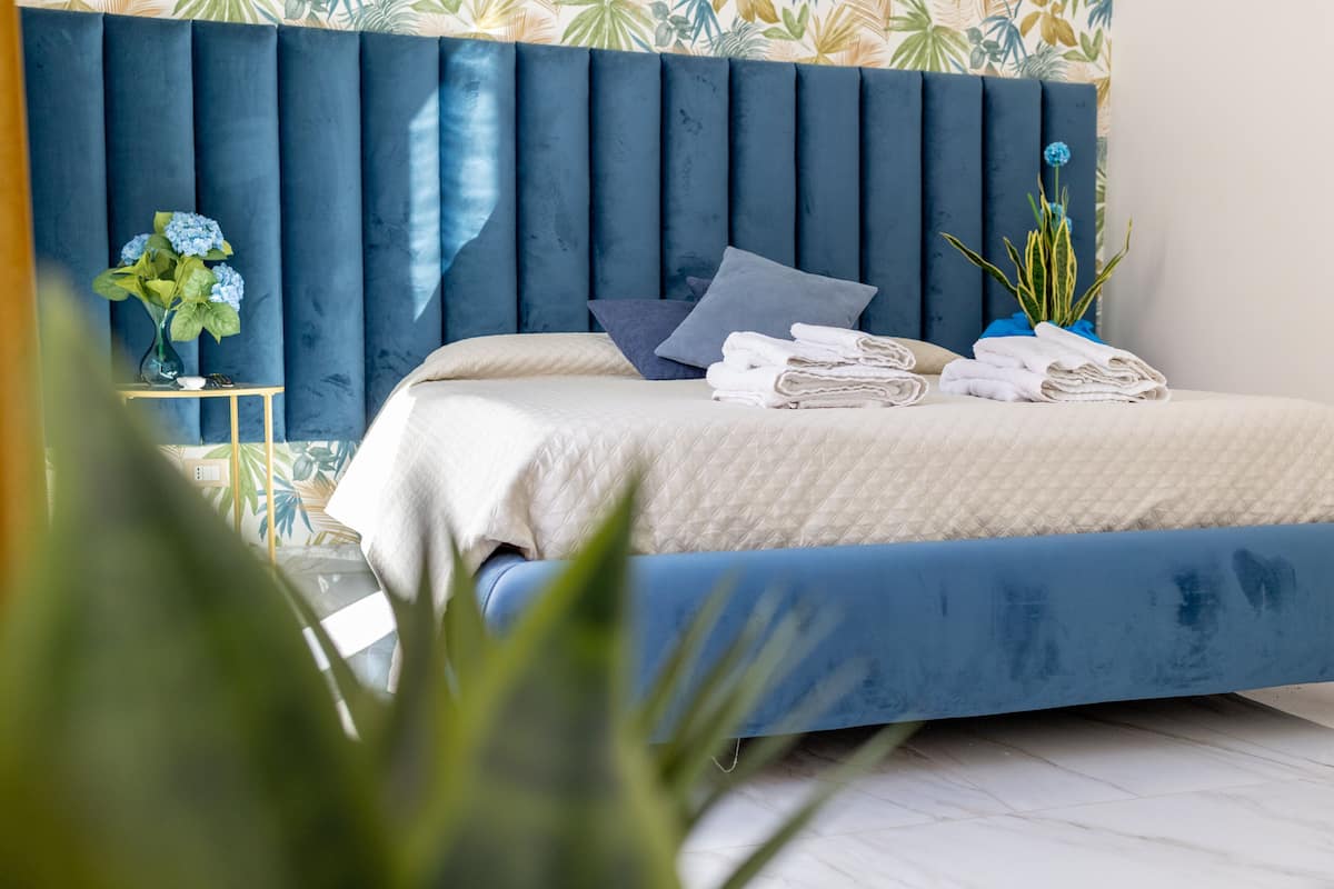 Quarto com cabeceira azul, cama com roupa de cama branca, mesinha de cabeceira com um vaso de flores e uma planta ao lado.