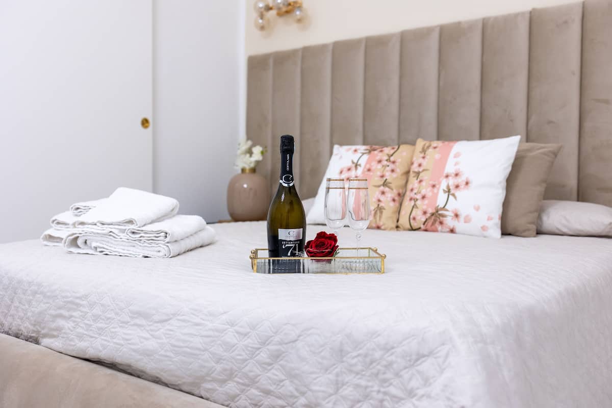 Uma cama bem arrumada com uma toalha branca dobrada, uma garrafa de vinho, dois copos e uma rosa em uma mesa de cabeceira.