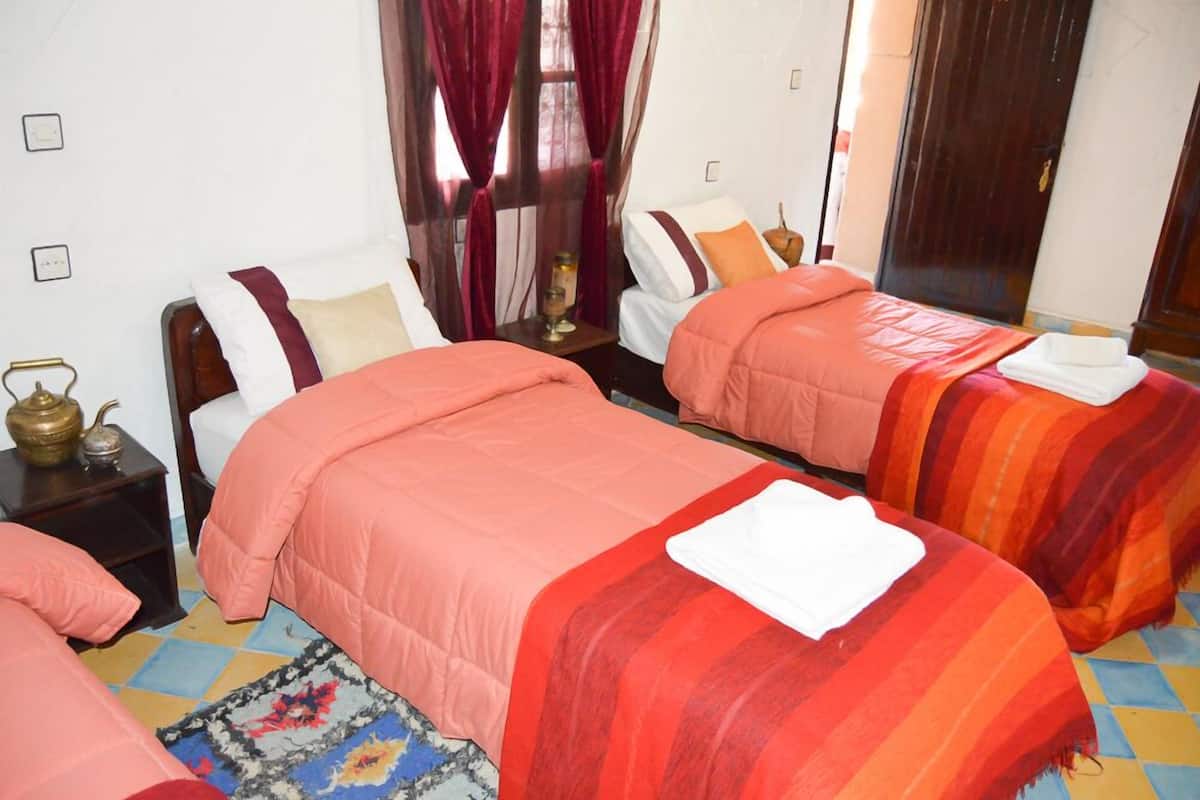 A hotel room with two beds, a wooden headboard, a small bedside table with a lamp, and a window with curtains.