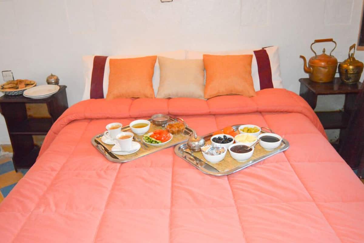 A bed with a pink blanket, two trays of breakfast items, and a nightstand with a teapot.