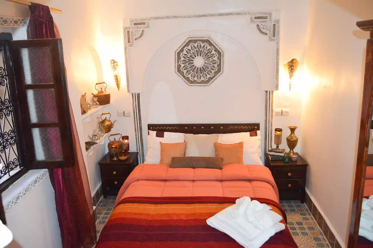 A hotel room with a bed, a nightstand with a lamp, a decorative wall piece, and a window with curtains.