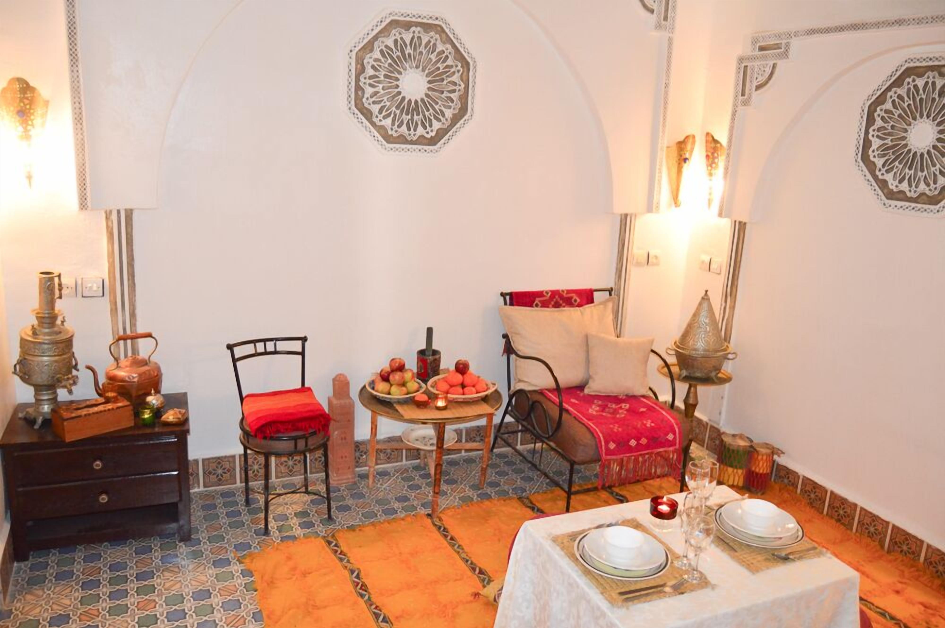 Um quarto aconchegante com área de jantar, uma mesa pequena com frutas, uma cadeira e um sofá com manta vermelha.
