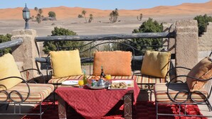 Une table à manger recouverte d’une nappe rouge, agrémentée de coussins à rayures, et offrant une vue sur le désert, avec ses dunes de sable et sa végétation clairsemée.