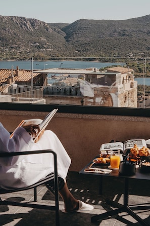 Una persona leyendo un libro en un balcón con una mesa de comida y bebidas, con vistas a un cuerpo de agua y edificios.