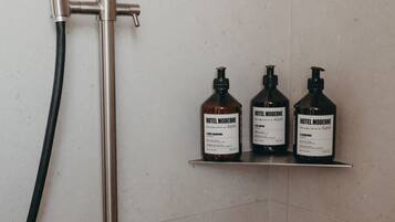 A shower with a handheld showerhead and a wall-mounted faucet. There are three bottles of Hotel Moderne body wash on a shelf.