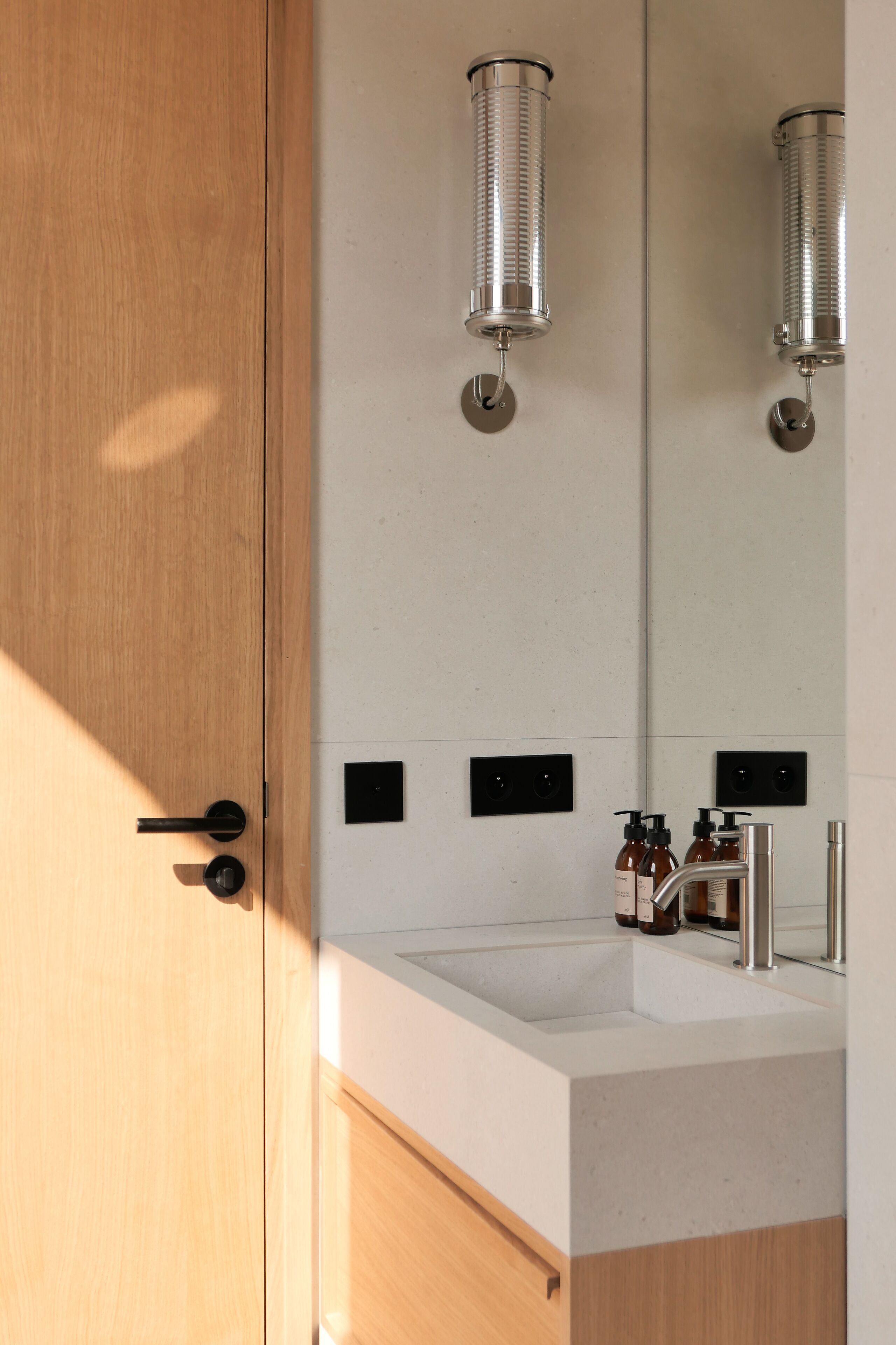 Ein modernes Badezimmer mit Holztür, Waschbecken in Weiß und zwei Wandleuchten.